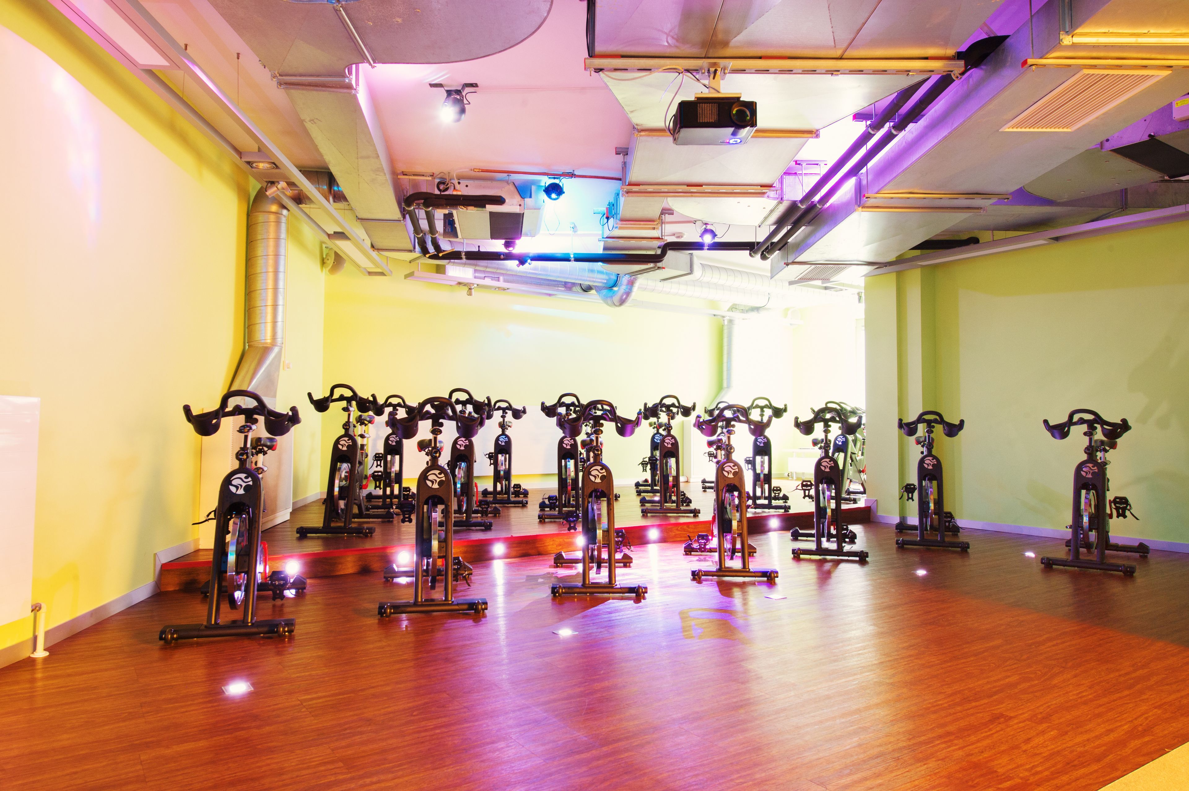 Uma academia com bicicletas ergométricas dispostas em fileiras sobre um piso de madeira, com iluminação e sistemas de ventilação no teto.