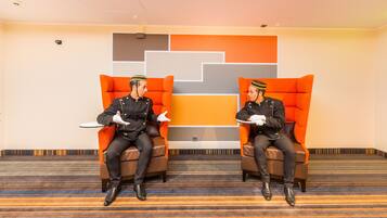 Twee mannen in uniform zitten op oranje stoelen in een kamer met een gestreept tapijt en een geometrisch wandontwerp.