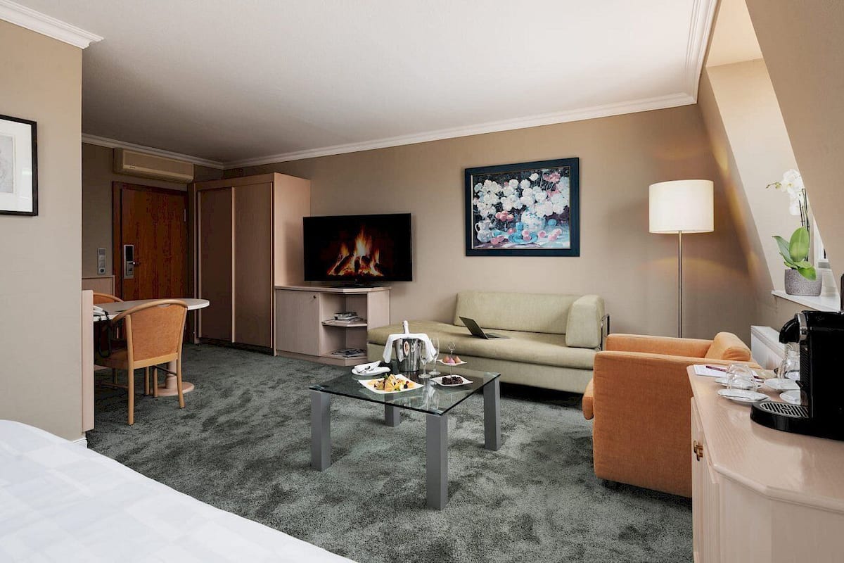 A hotel room with a sofa, armchair, coffee table, TV, and a painting on the wall.