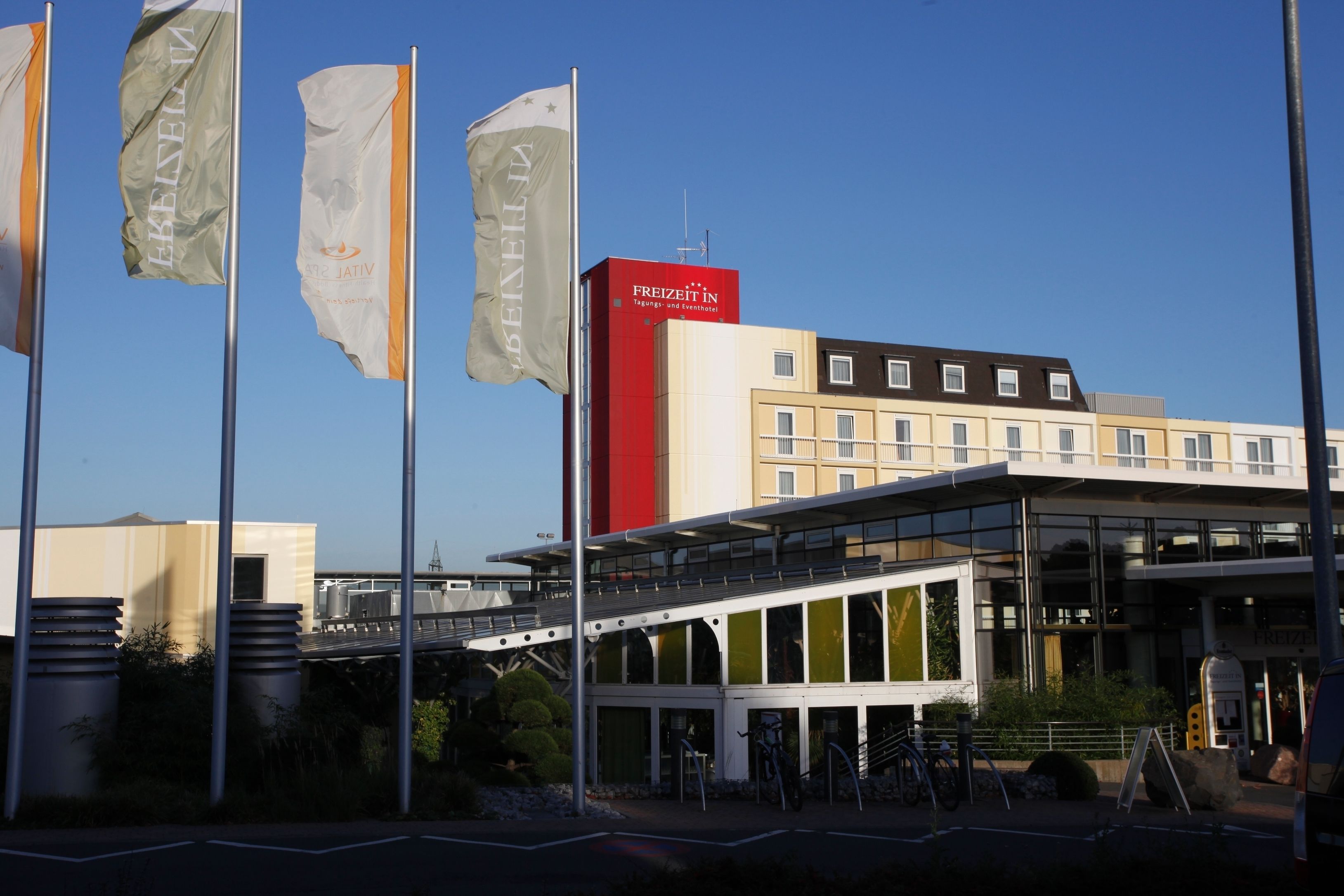 Un bâtiment moderne à la façade rouge et blanche, orné de plusieurs drapeaux et doté d’une galerie couverte.