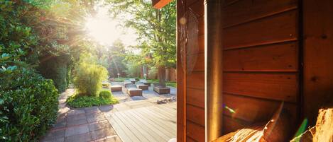 Una terraza de madera con mobiliario exterior, rodeada de follaje y un edificio de madera.