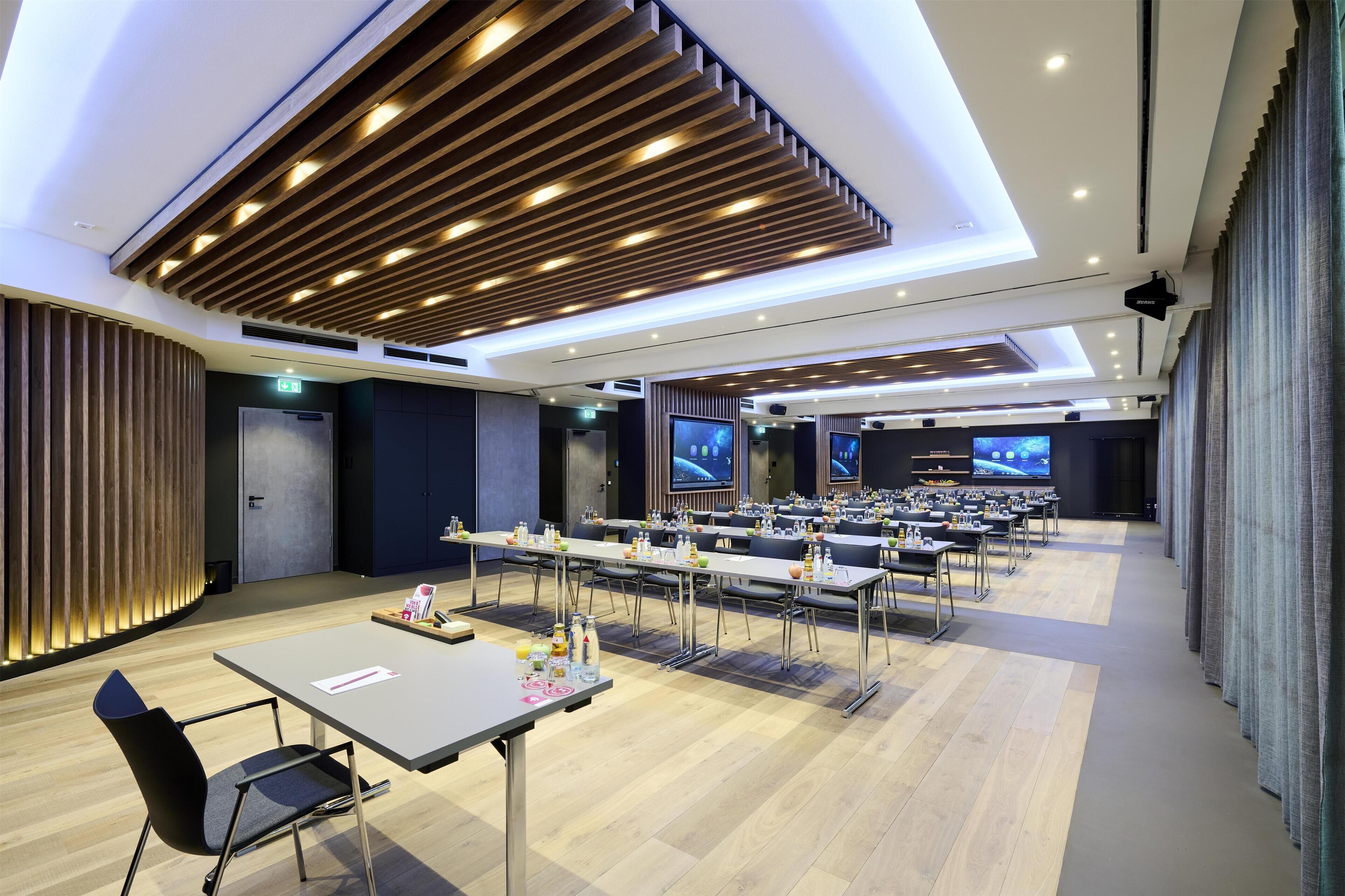 Une salle de conférence moderne, dotée de longues tables, de chaises et d’une scène avec des écrans.
