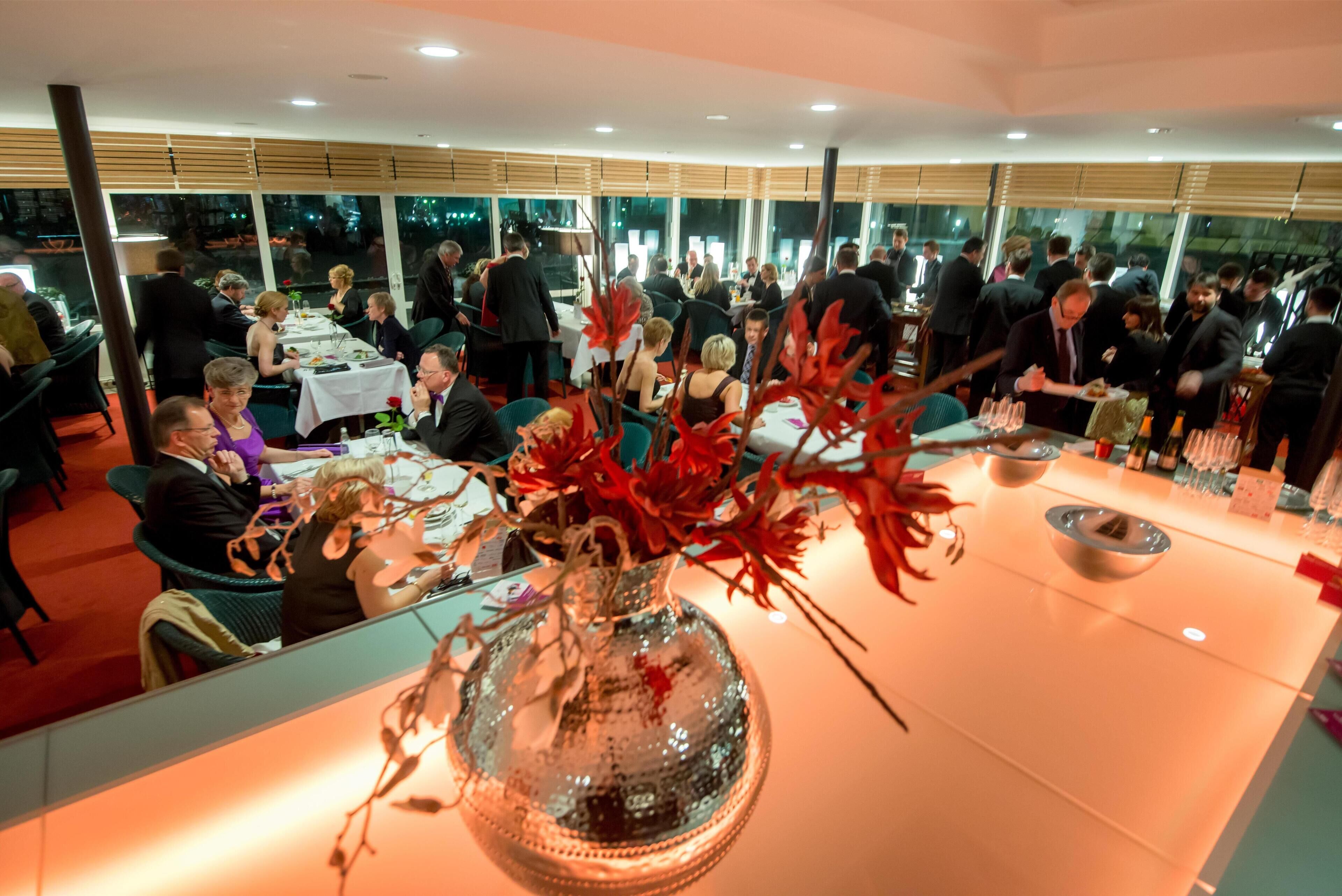 Un restaurant avec des tables rondes, des clients qui dînent et un vase décoratif en verre contenant des fleurs rouges.