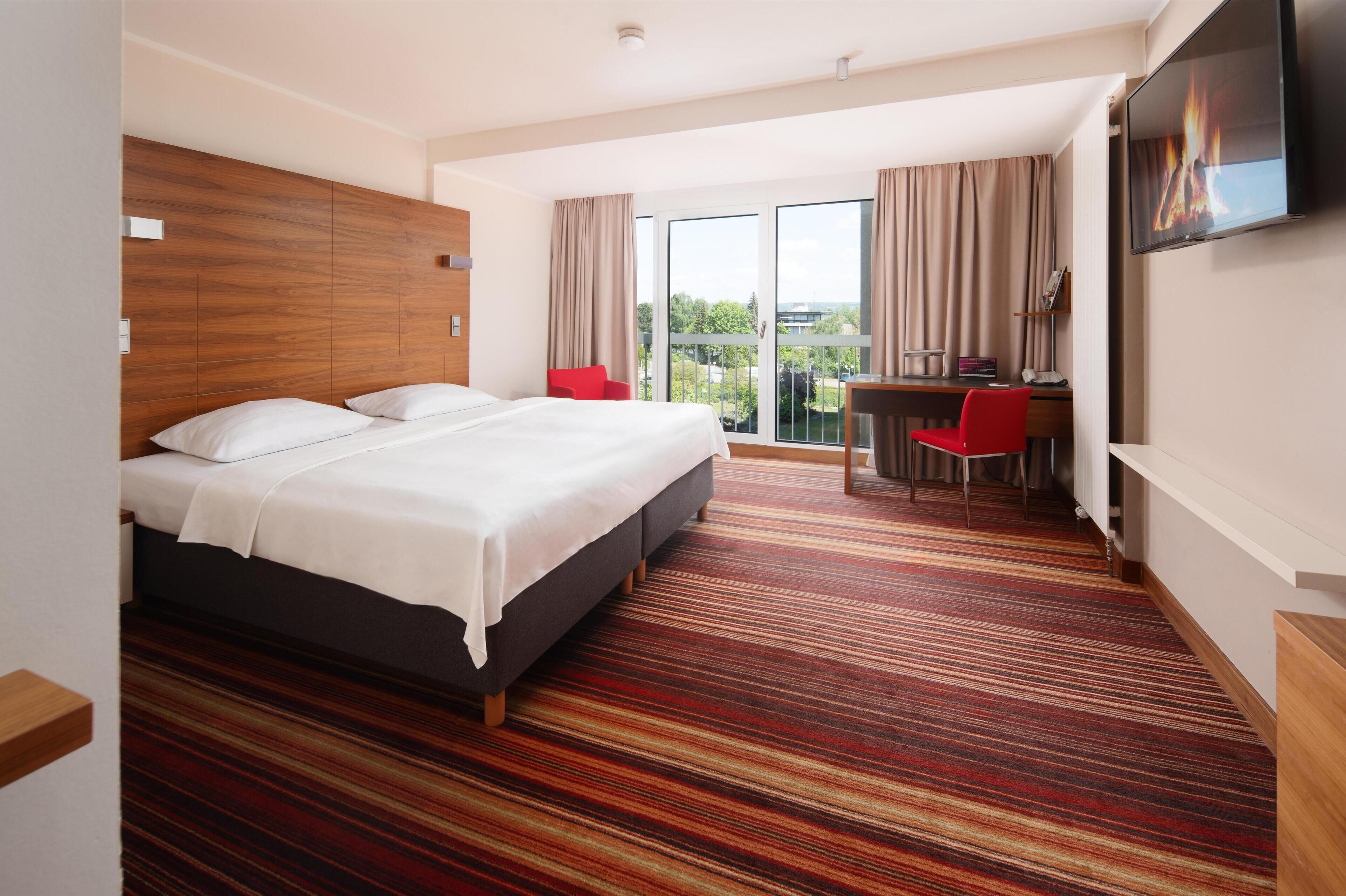 Una habitación de hotel con una cama grande, un escritorio con silla, un televisor y un balcón con cortinas.