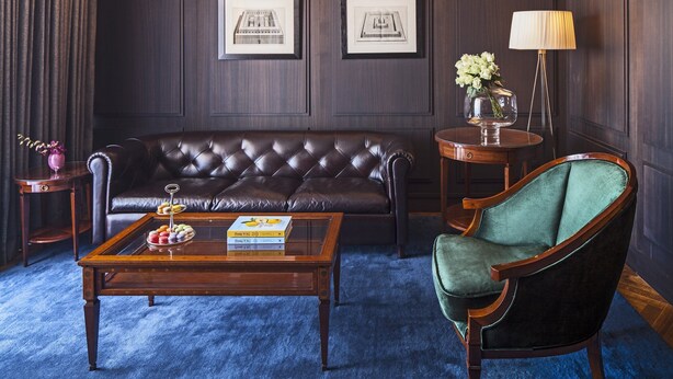 A dark-themed living room with a leather sofa, a green armchair, a wooden coffee table with books and a fruit tray, a side table with a vase of flowers, and wall-mounted picture frames. - The David Citadel Jerusalem (Jerusalem)
