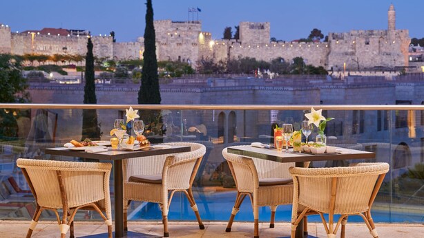 A terrace with wicker chairs and tables set for dining, overlooking a historic stone wall and a body of water. - The David Citadel Jerusalem (Jerusalem)