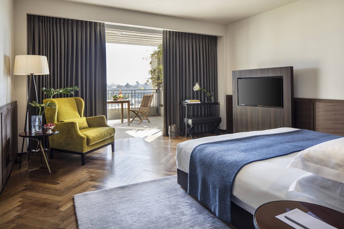 Quarto de hotel com cama, sofá, televisão, varanda e vista, além de mesa com luminária e planta.
