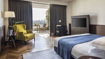 Habitación de hotel con cama, sofá, televisor, balcón con vistas y una mesa con lámpara y planta.