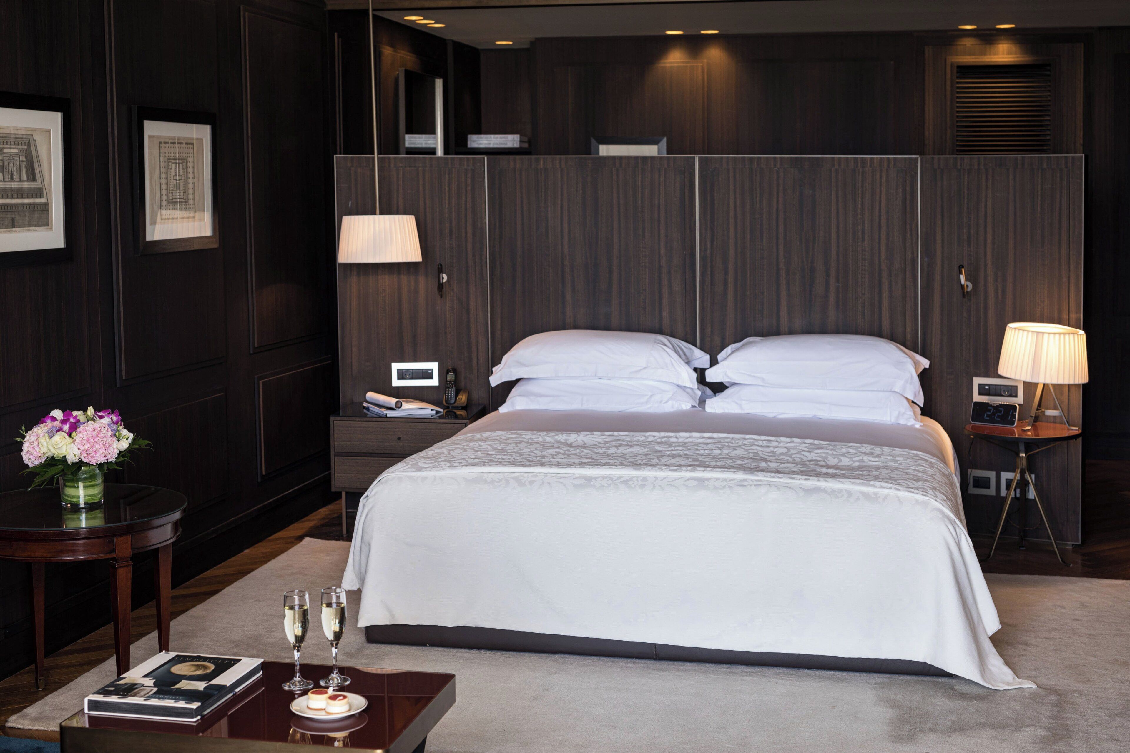Una habitación de hotel moderna con una cama grande, una mesita de noche con una lámpara, un buró con un teléfono y una pequeña mesa con un jarrón de flores y un libro.