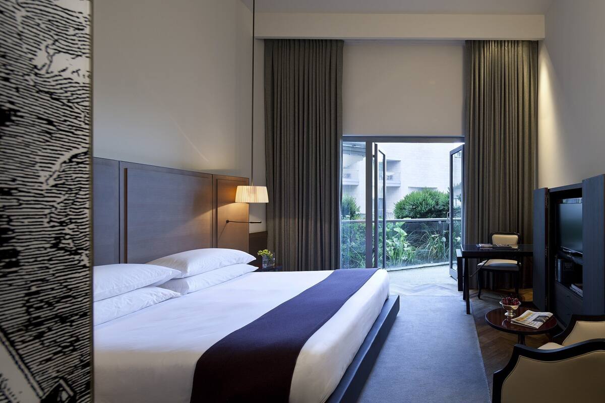 Quarto de hotel com cama grande, televisão, escrivaninha com cadeira e varanda com cortinas.