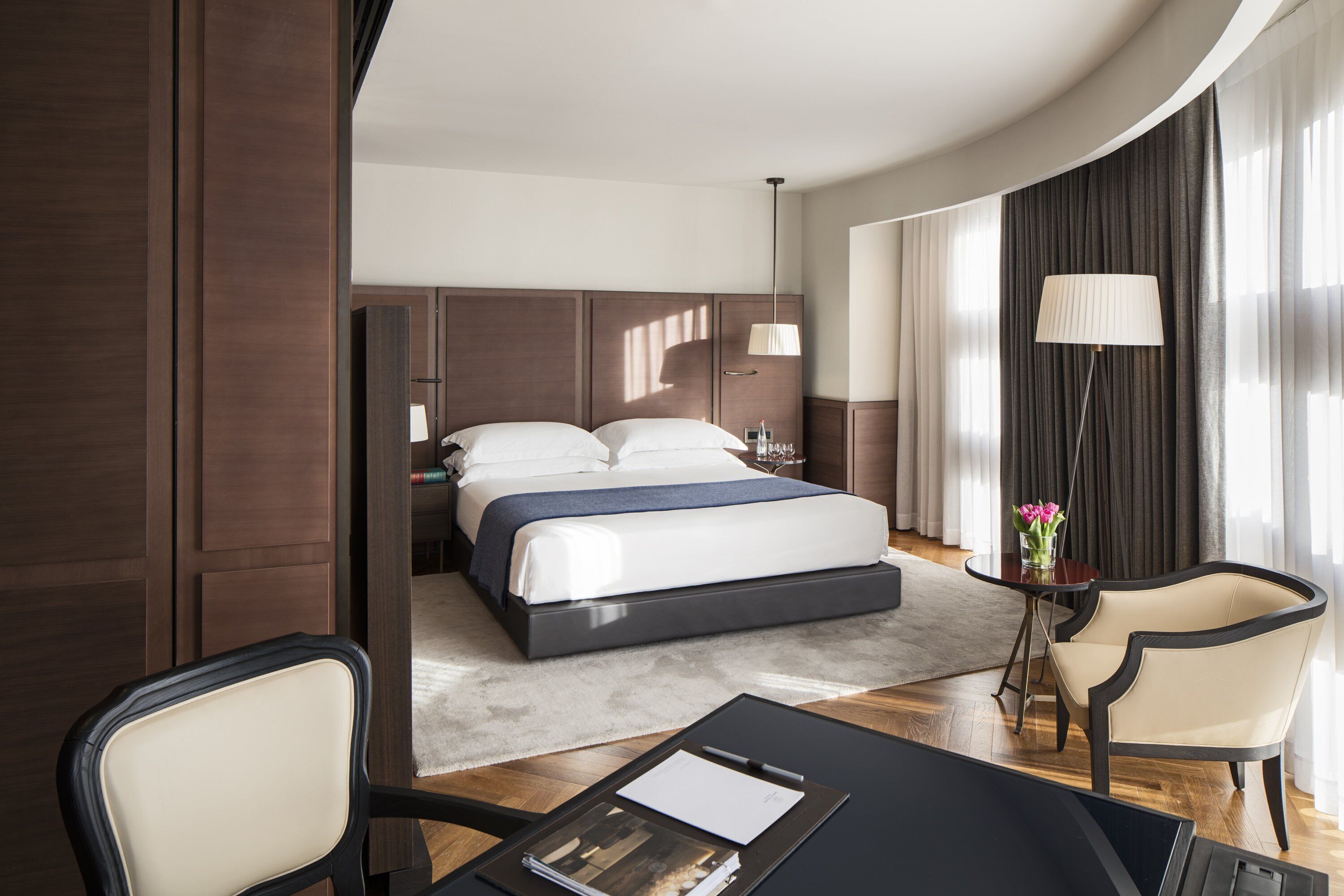 A modern hotel room with a large bed, a desk with a chair, a lamp, and a view of the outdoors.