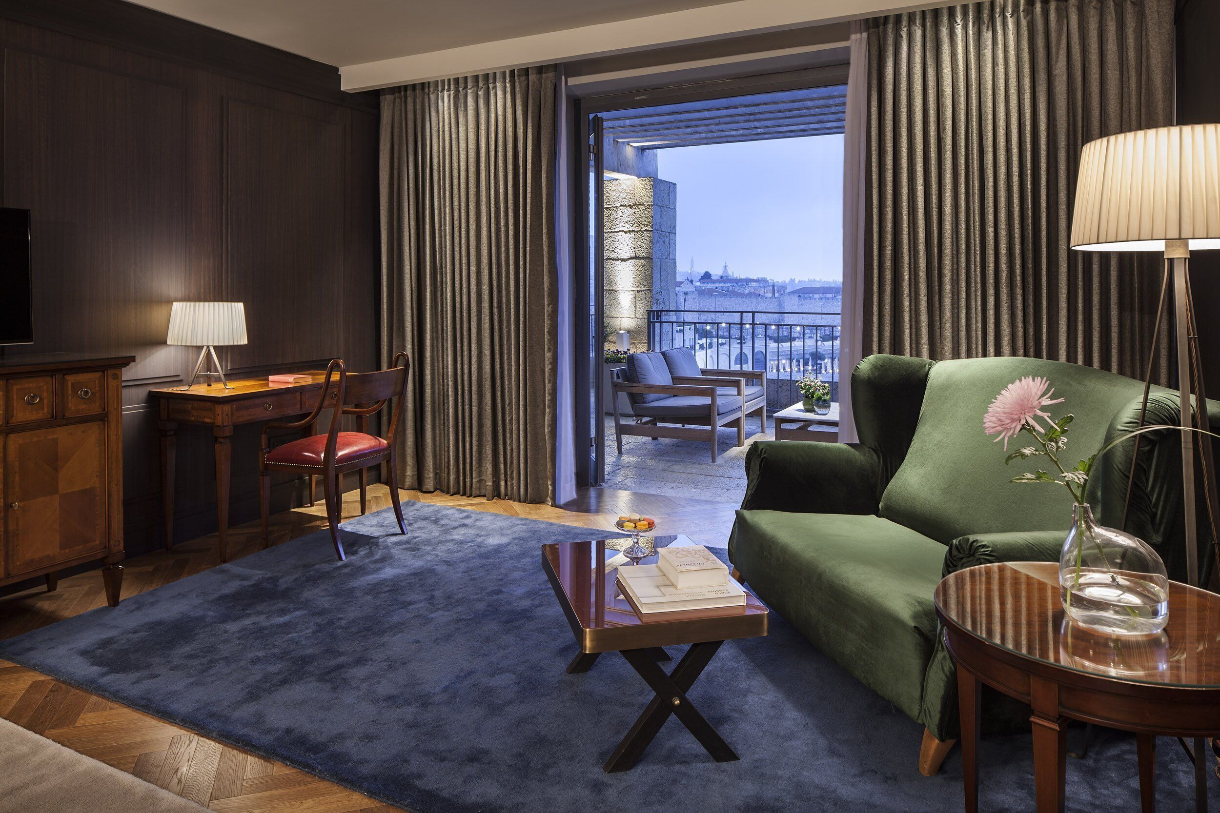 Habitación de hotel con balcón, un sofá verde, una mesa de centro de madera, un escritorio con lámpara y un televisor.