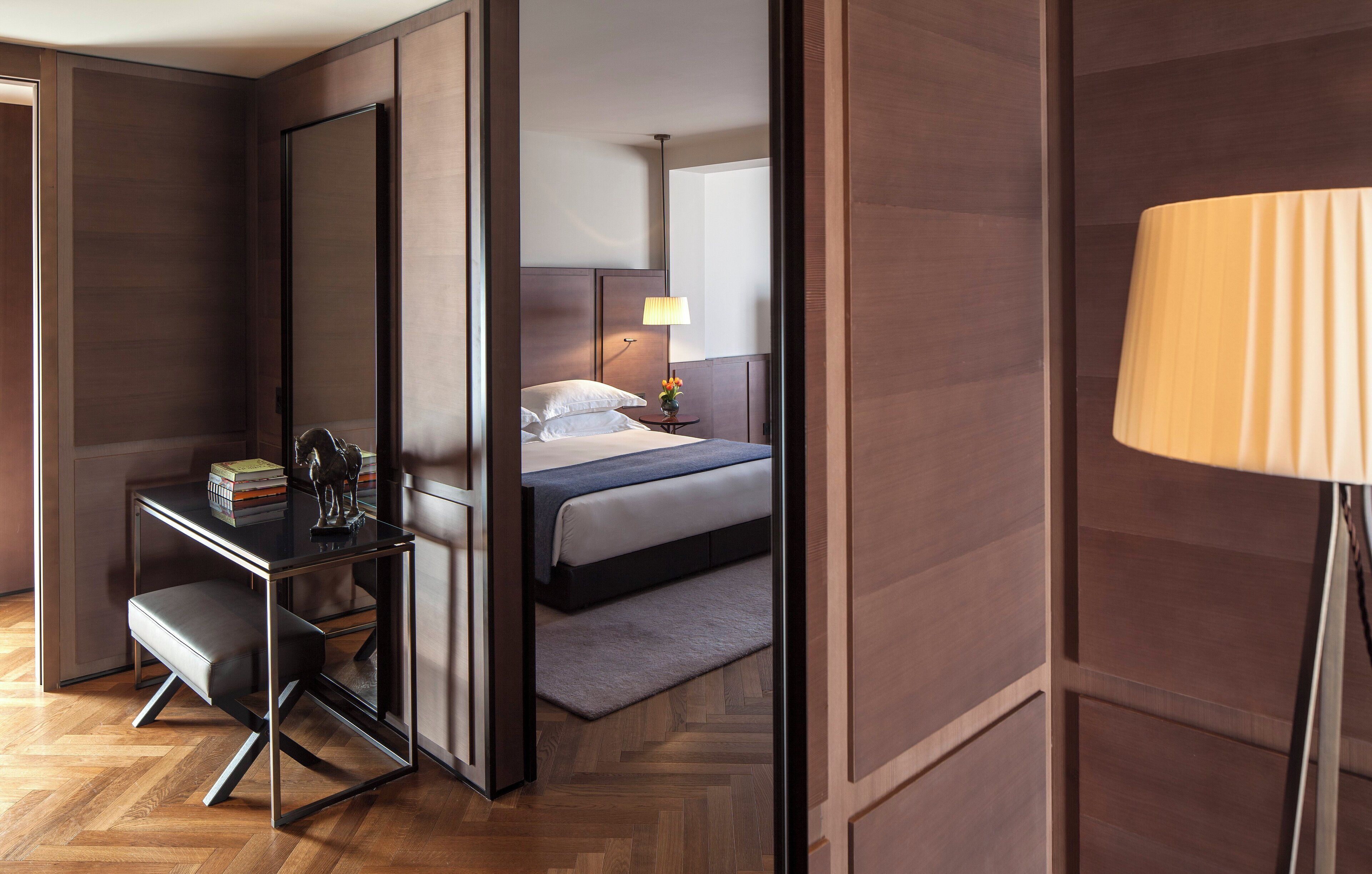 A modern hotel room with a wooden floor, a desk with a chair, a bed with a pillow, a nightstand with a lamp, and a large mirror.