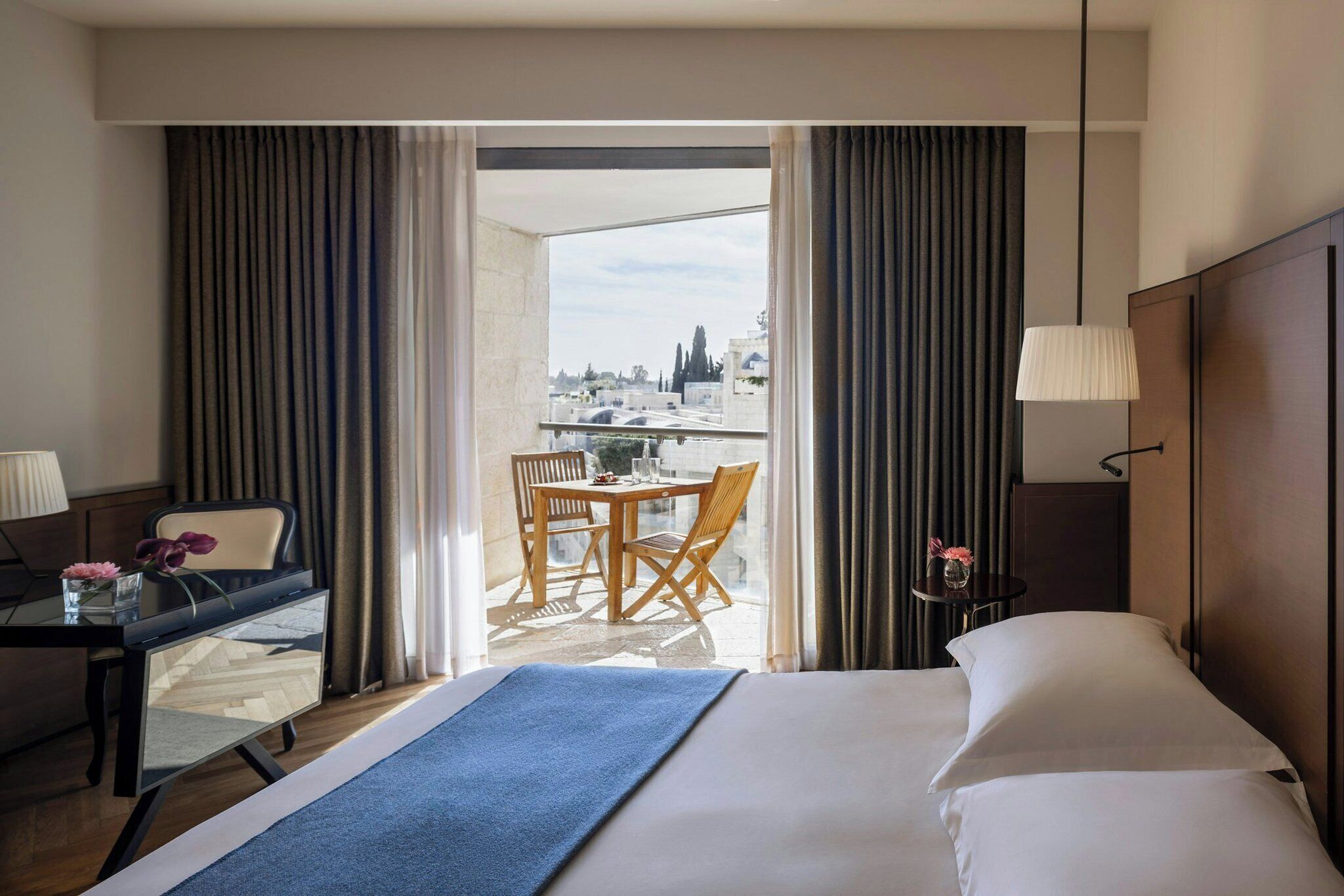 Habitación de hotel con cama, escritorio, silla, mesa y vistas a la ciudad a través de amplios ventanales.