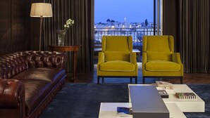 A modern living room with a leather sofa, two yellow armchairs, a coffee table, and a lamp. A view of a cityscape is visible through a large window. - The David Citadel Jerusalem (Jerusalem)