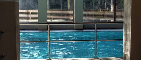 Zona de piscina con suelo de baldosas, barandillas de metal y grandes ventanales con vistas a una terraza de madera.