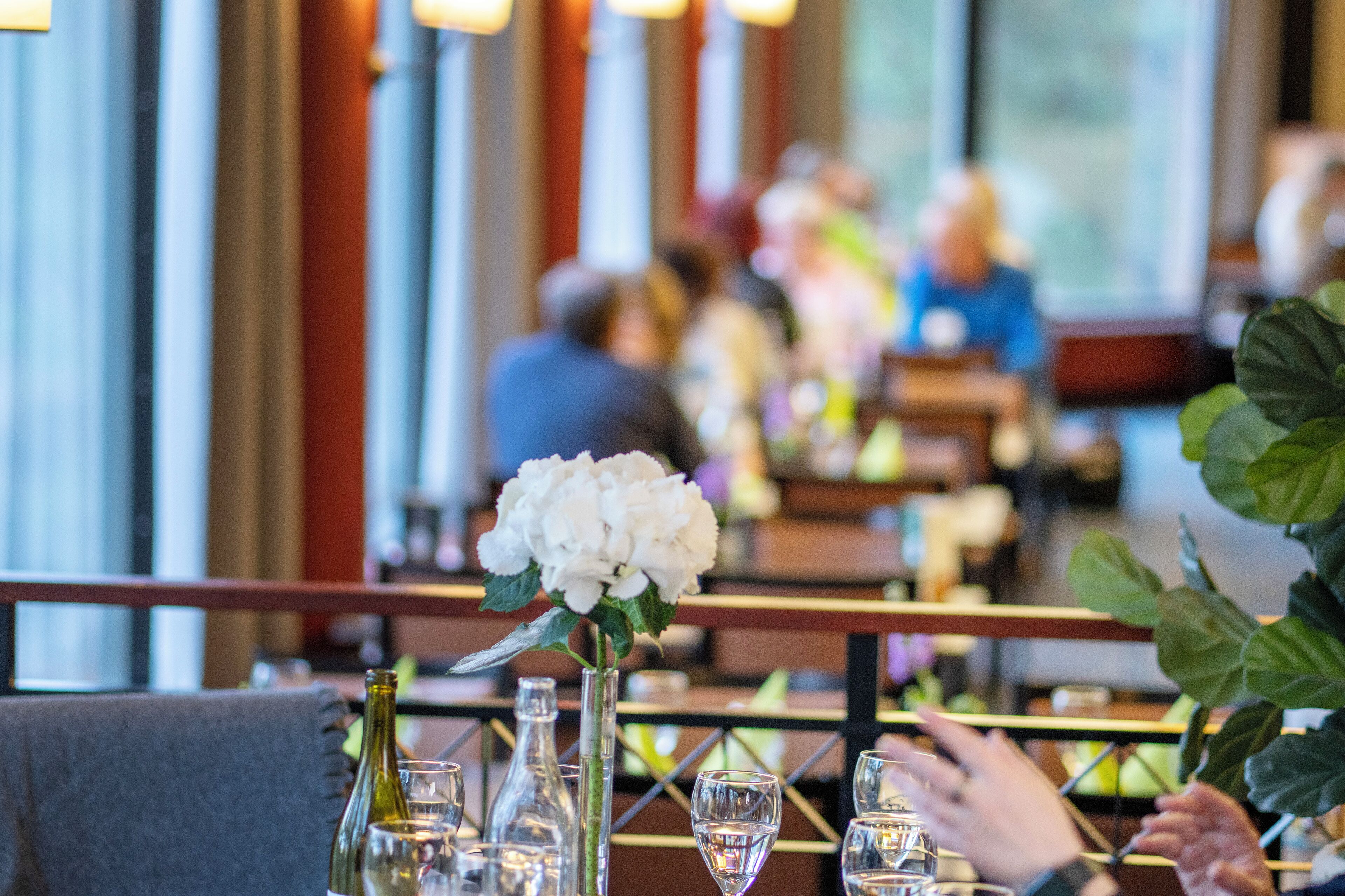 A restaurant interior with tables set for dining, featuring clear glassware and a vase with white flowers.
