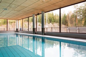 En inomhuspool med väggar av genomskinligt glas, stora fönster och utsikt över ett skogsområde.