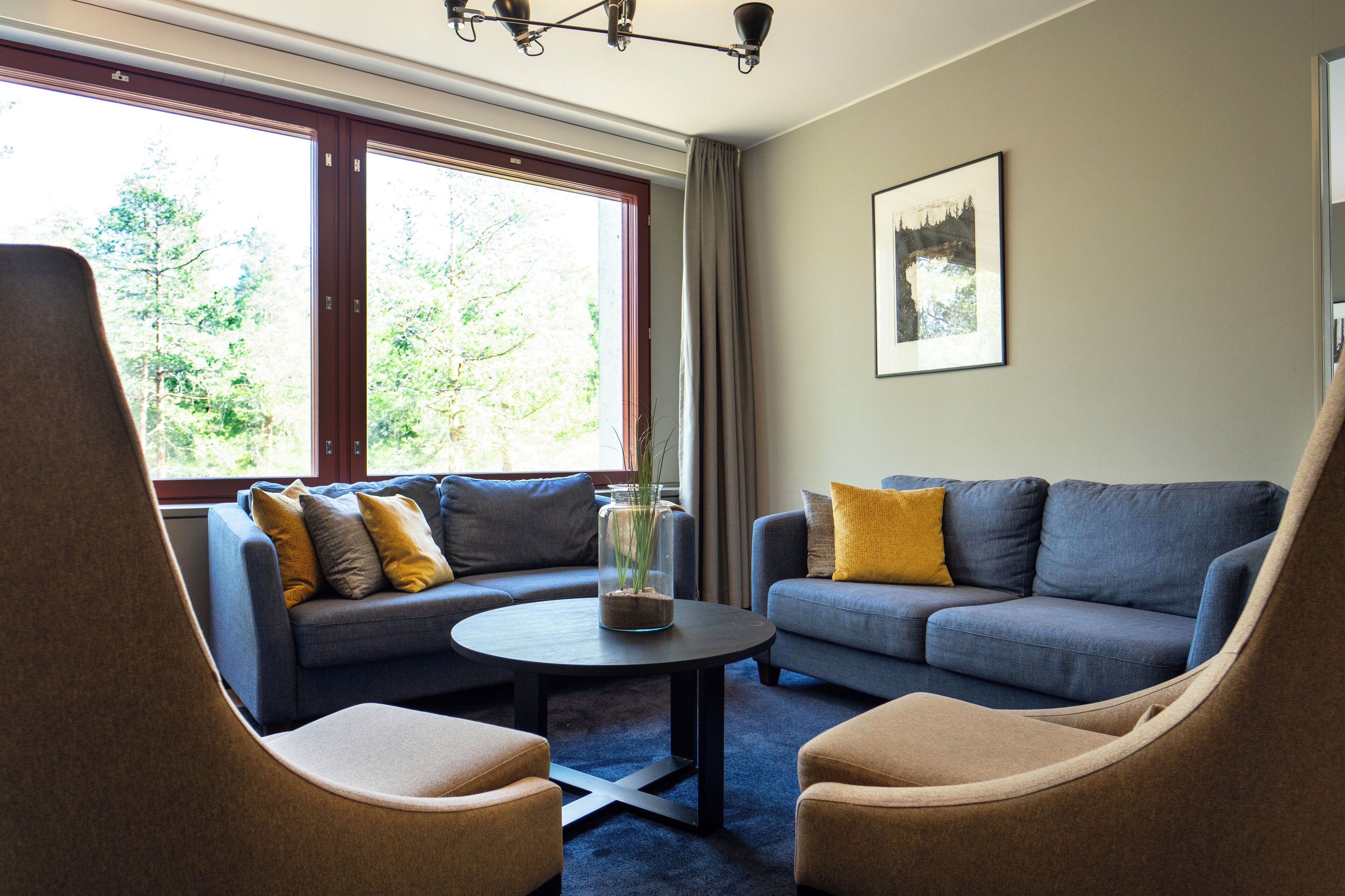 Ein modernes Wohnzimmer mit einer Couch, zwei Sesseln, einem Couchtisch und einem großen Fenster mit Blick auf Bäume.