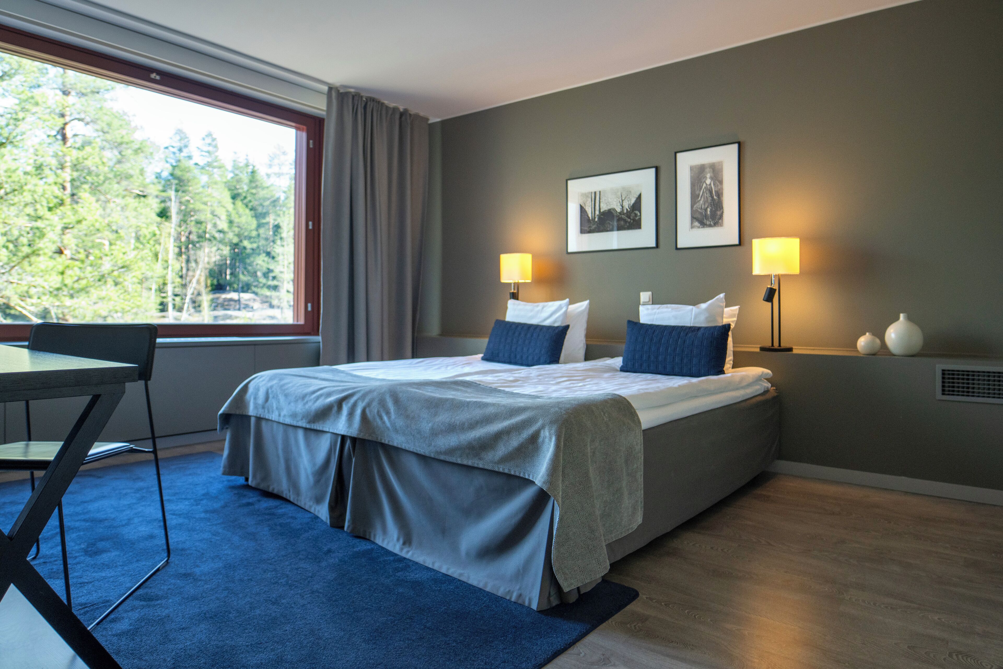 Una habitación de hotel moderna con una cama grande, dos lámparas de noche, un escritorio y vistas a árboles por la ventana.