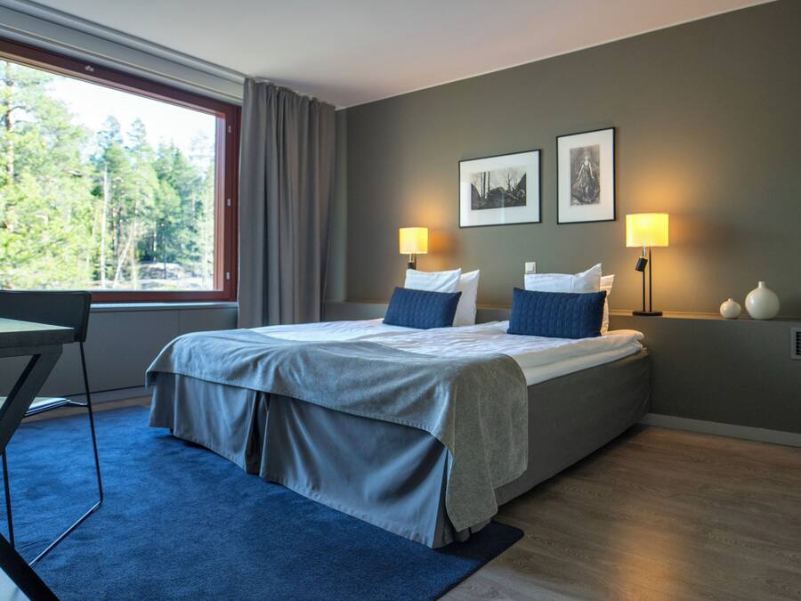 A modern hotel room with a large bed, two bedside lamps, a desk, and a view of trees through a window.