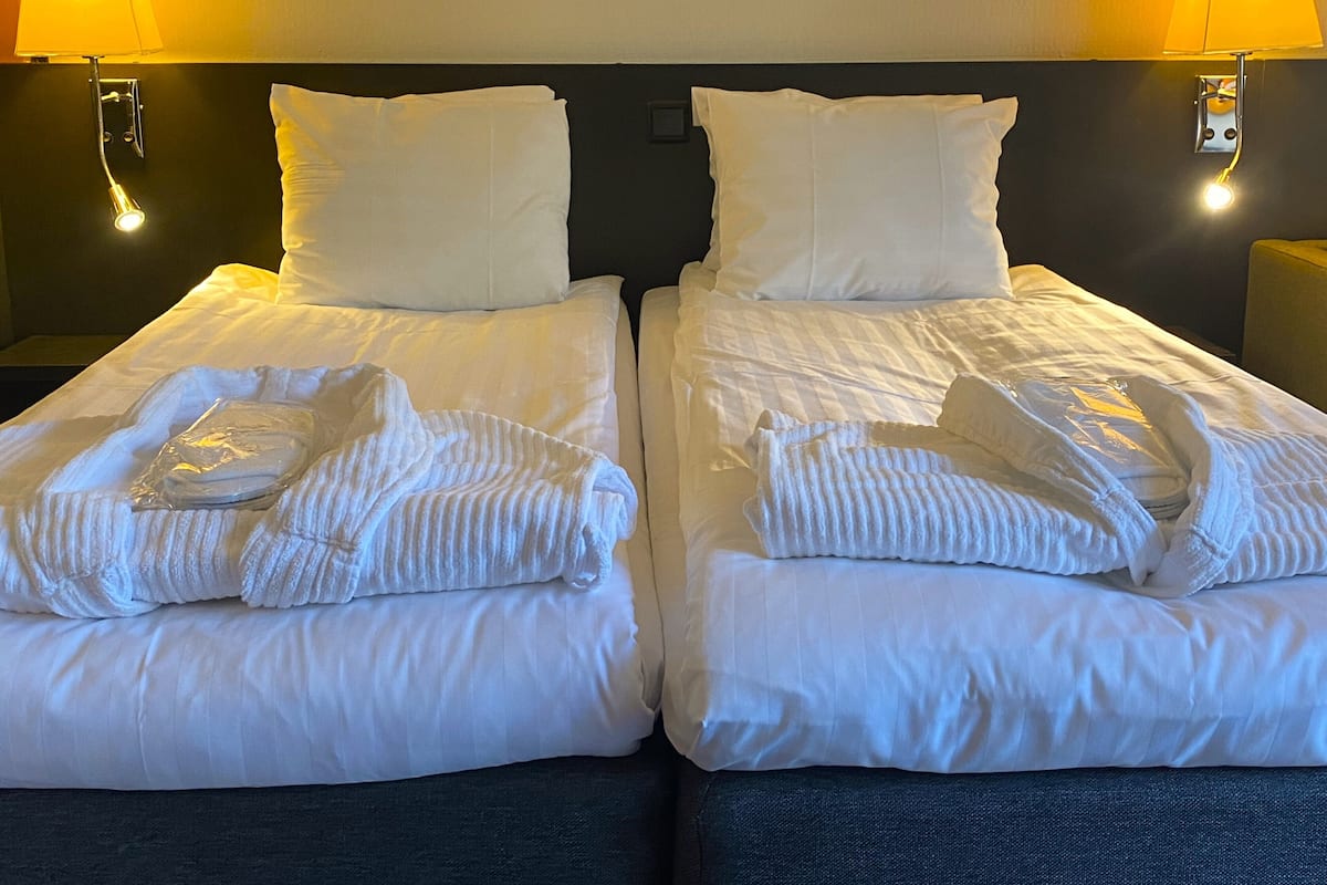 Hotellihuone, jossa on kaksi sänkyä, molemmissa valkoiset lakanat ja tyynyt, sekä kaksi yöpöytävalaisinta.
