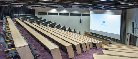 Un salón de conferencias con filas de escritorios y sillas de madera, una pantalla de proyección grande y un suelo cubierto de moqueta púrpura.