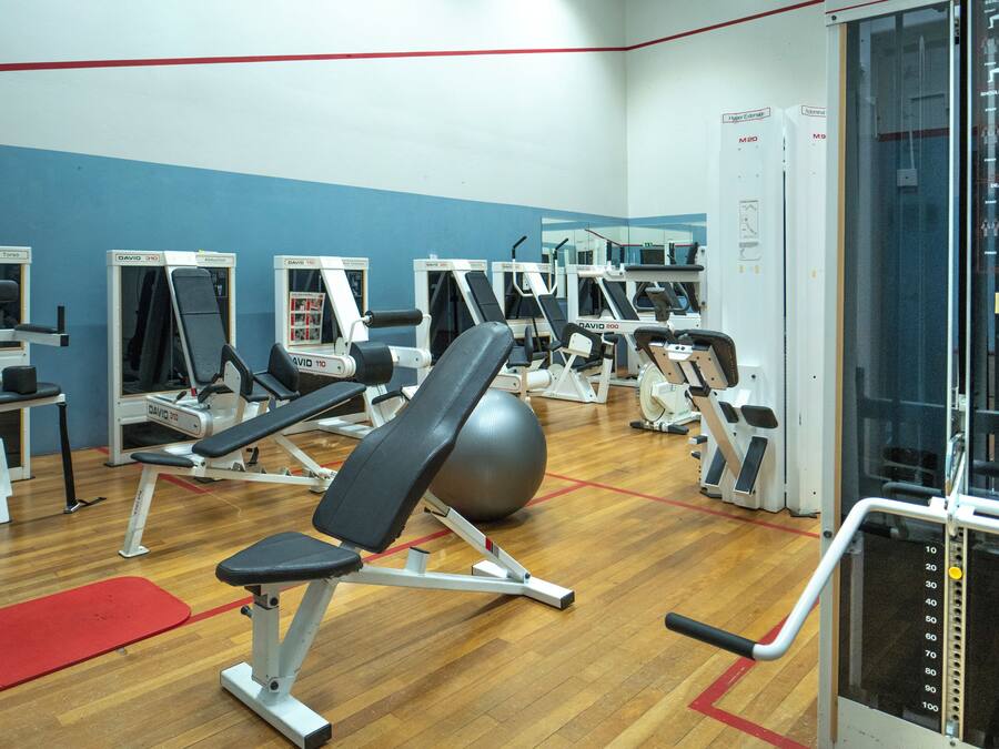 A gym with various exercise machines, a red mat, and a wooden floor.