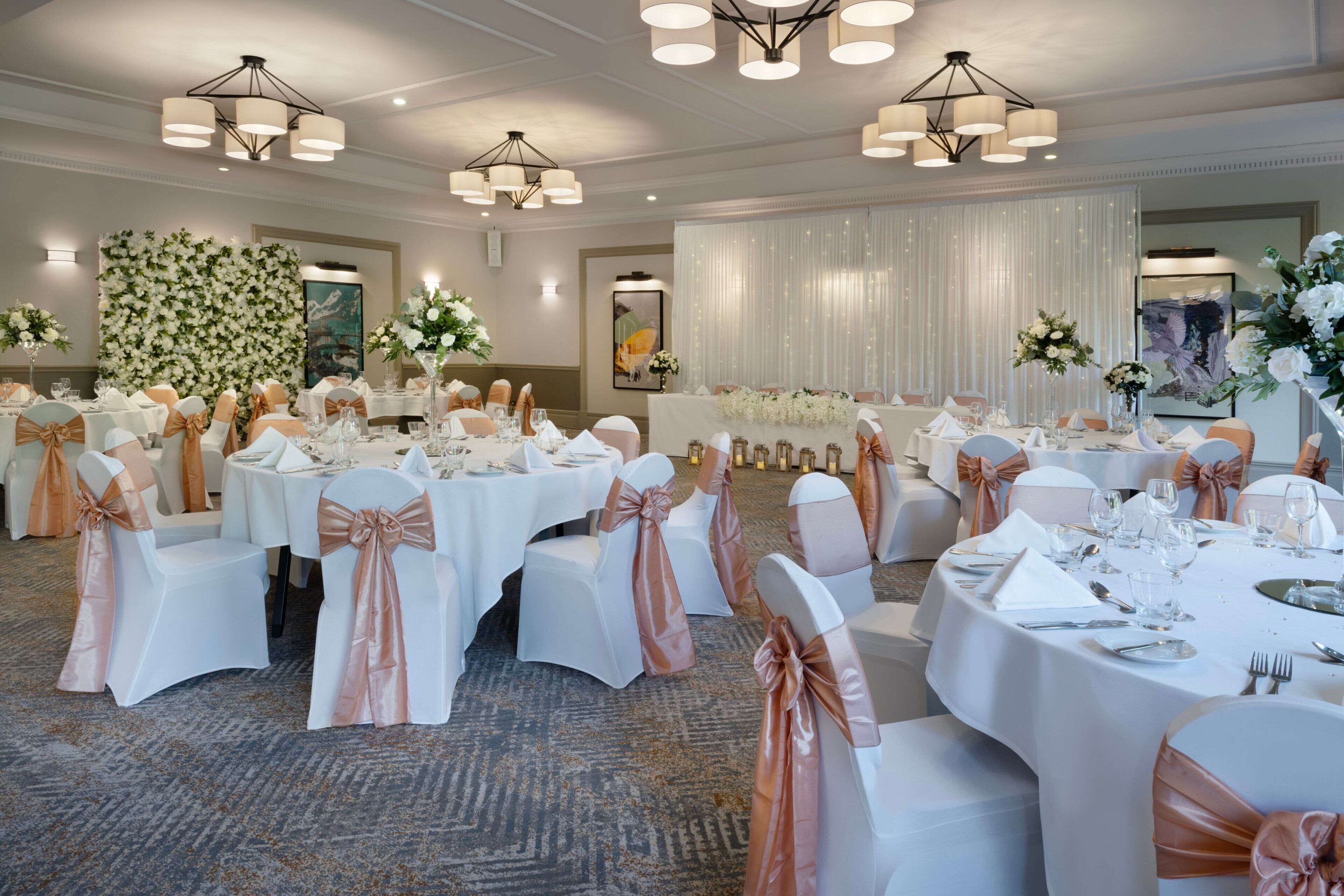 Een feestzaal met ronde tafels, witte stoelen met roze gouden linten, en kroonluchters.