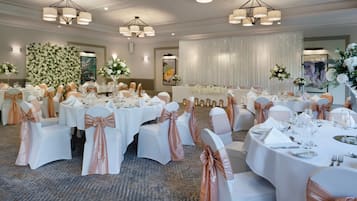 Een feestzaal met ronde tafels, witte stoelen met roze gouden linten, en kroonluchters.