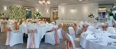 A banquet hall with round tables, white chairs with rose gold sashes, and chandeliers.
