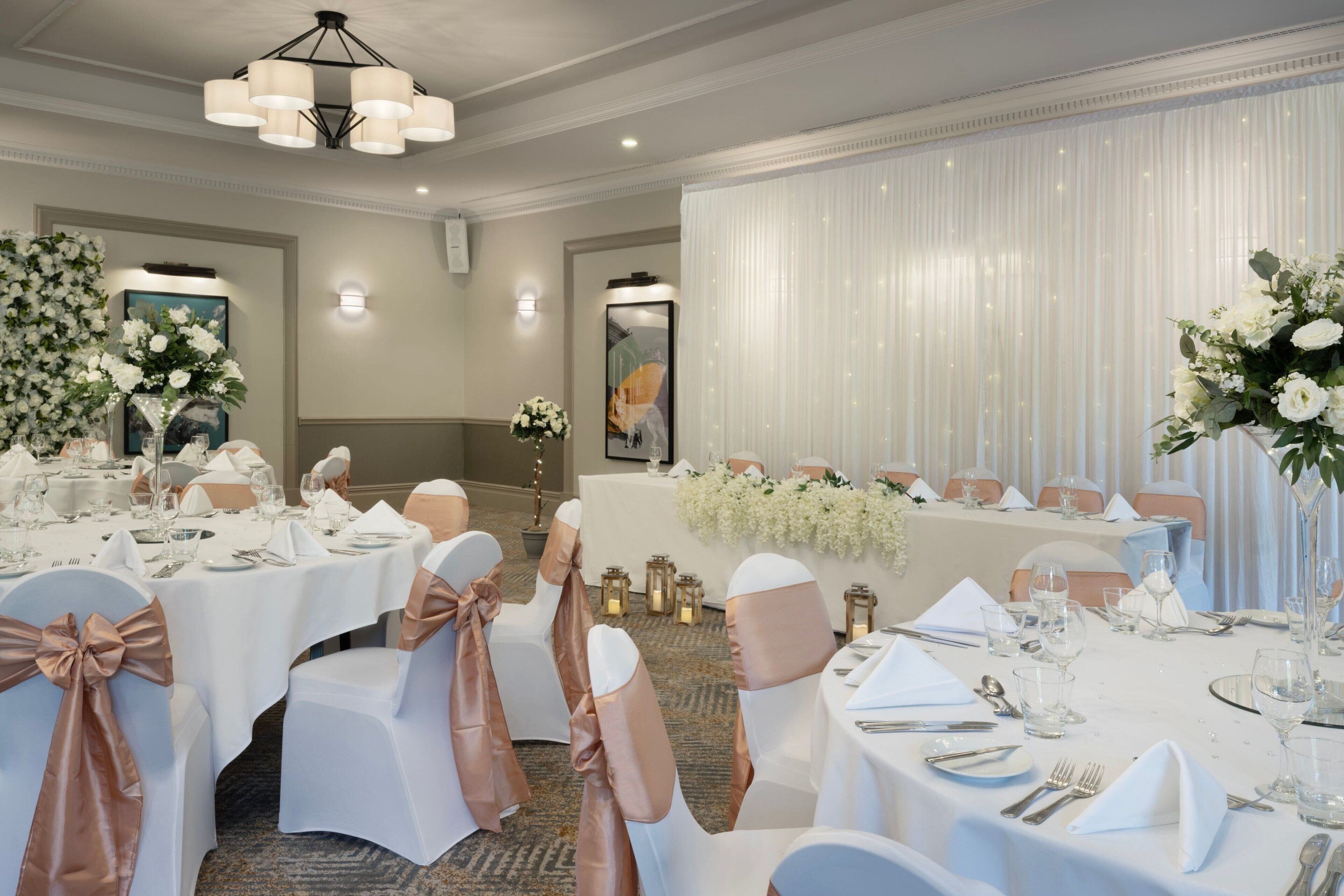 Un salón de banquetes con mesas redondas dispuestas para un evento formal, sillas blancas con cintas color rosa oro y centros de mesa florales.