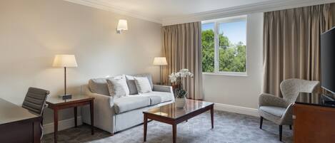 Habitación de hotel con sofá, dos sillones, una mesa de centro, televisión y ventana con cortinas.