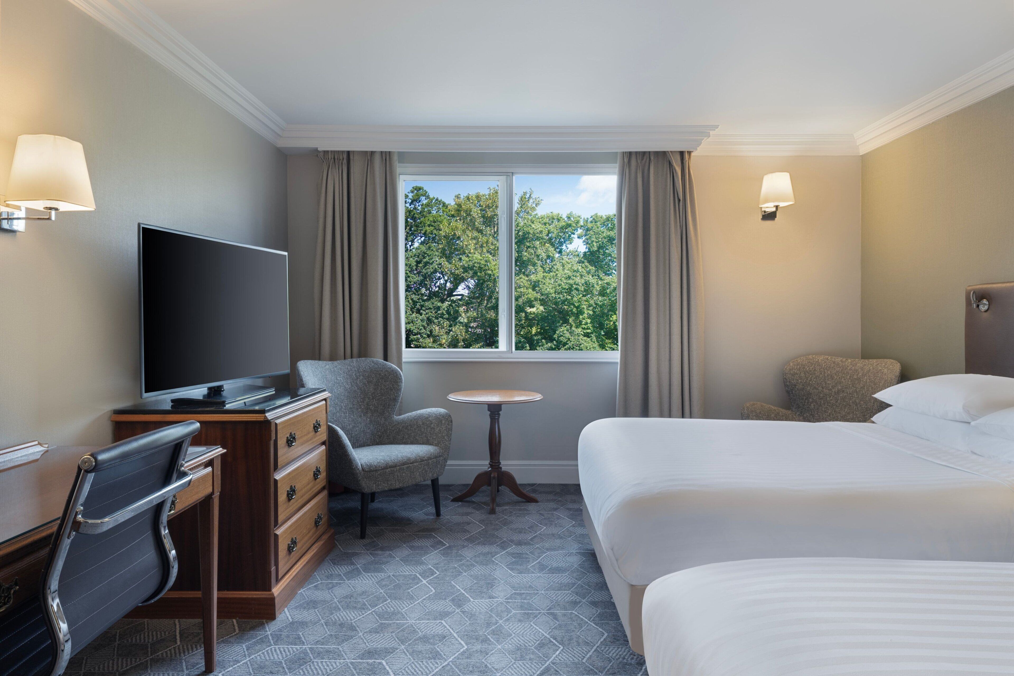 Ein Hotelzimmer mit einem Bett, einem Schreibtisch mit Fernseher, einem Sessel, einem kleinen Tisch und einem Fenster mit Vorhängen.