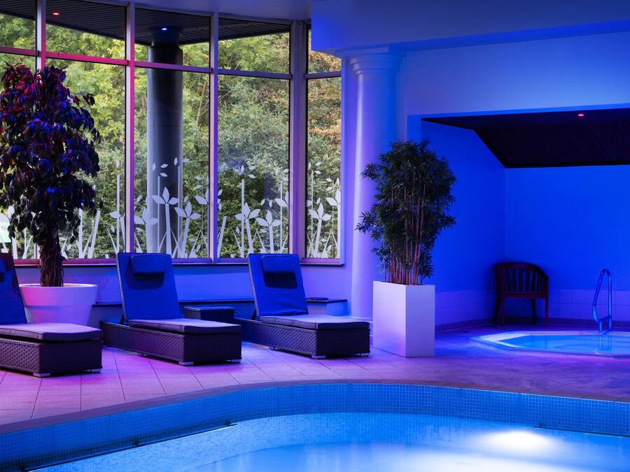 A modern indoor pool area with lounge chairs and a hot tub, surrounded by large windows and potted plants.