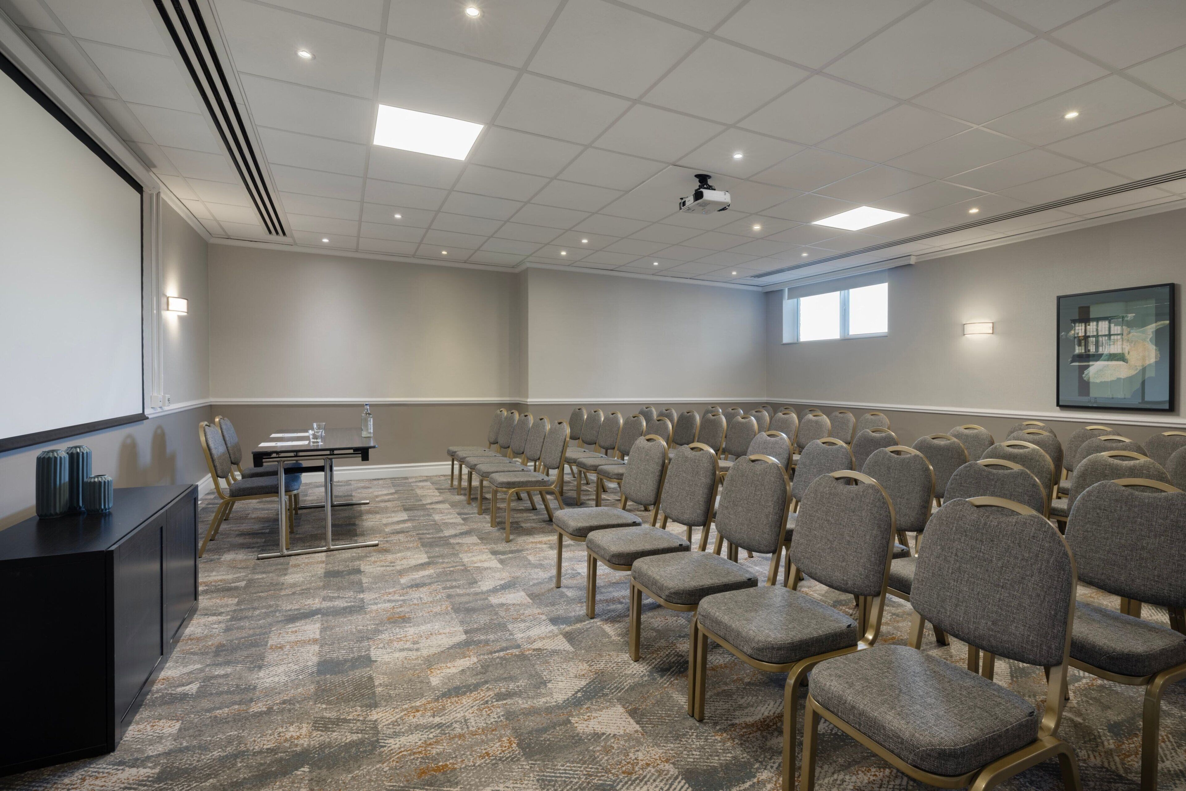 Une salle de conférence avec des rangées de chaises, un écran de projection et un sol recouvert de moquette.