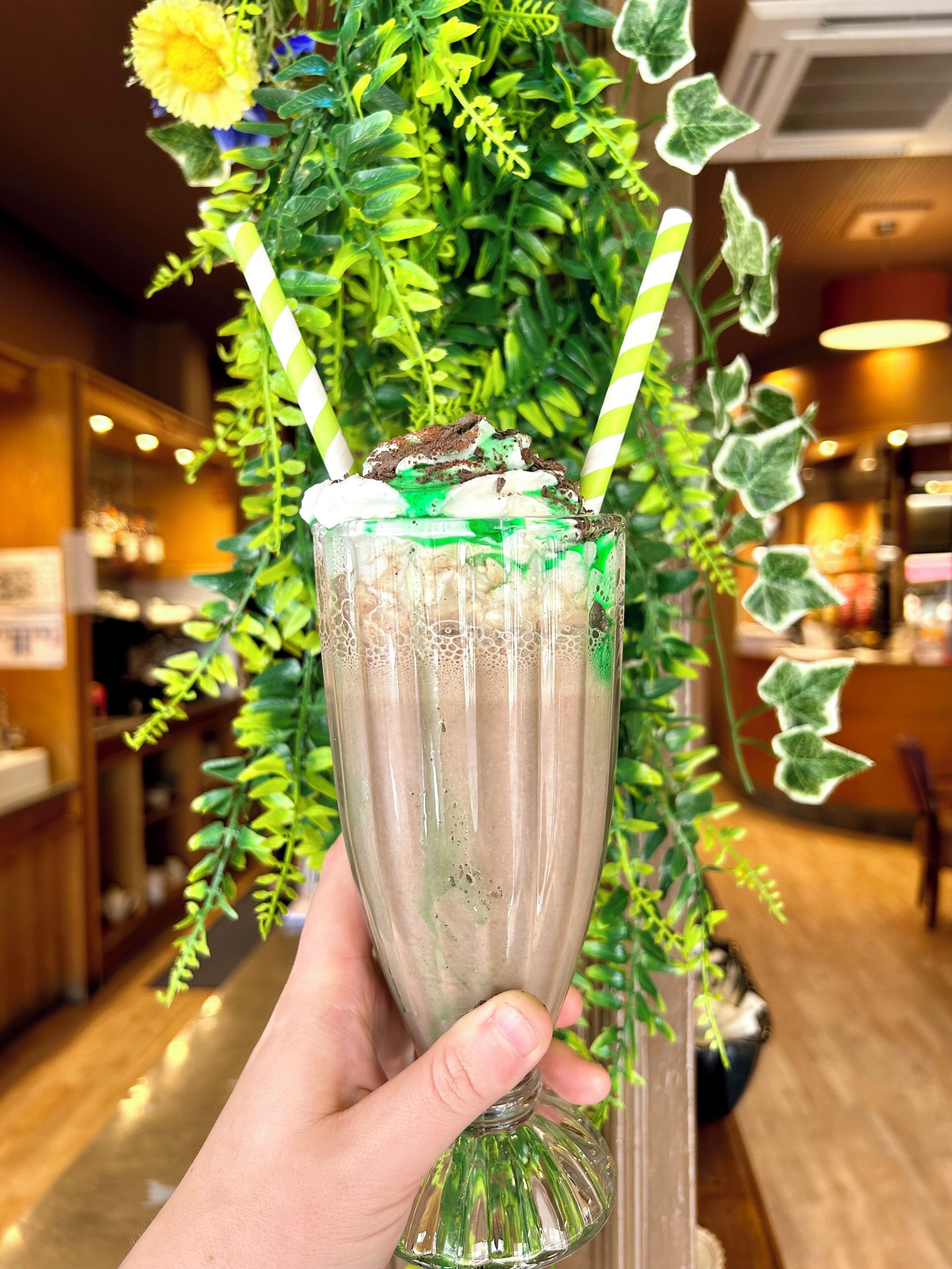 Une main tenant un verre contenant une boisson à plusieurs couches, entourée de verdure et éclairée par une lumière intérieure.
