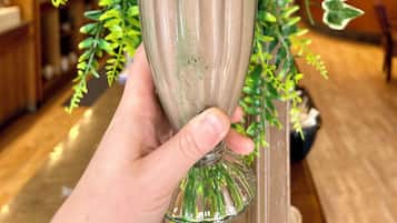 Una mano sosteniendo un vaso con una bebida en capas, rodeado de follaje e iluminación interior.