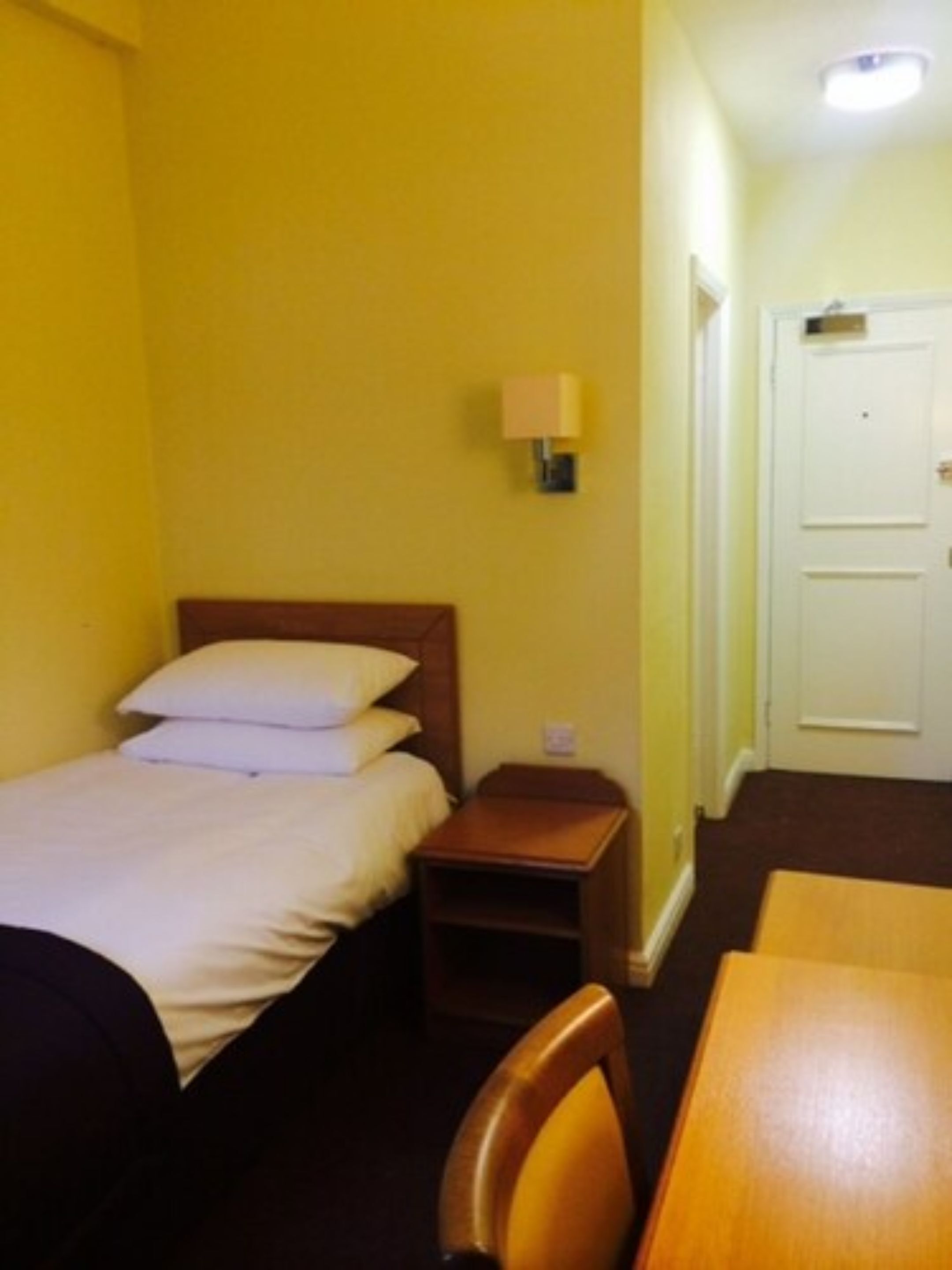 A hotel room with a bed, bedside table, lamp, and a wooden chair.