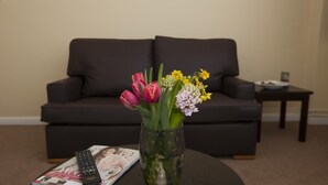 Una sala de estar con un sofá de color oscuro, un florero de vidrio con flores, un control remoto y una revista sobre una mesa de centro.