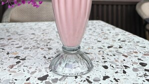 Un batido rosa con pajita verde y blanca a rayas, cubierto de crema batida y una cereza, sobre una mesa con superficie de terrazo.