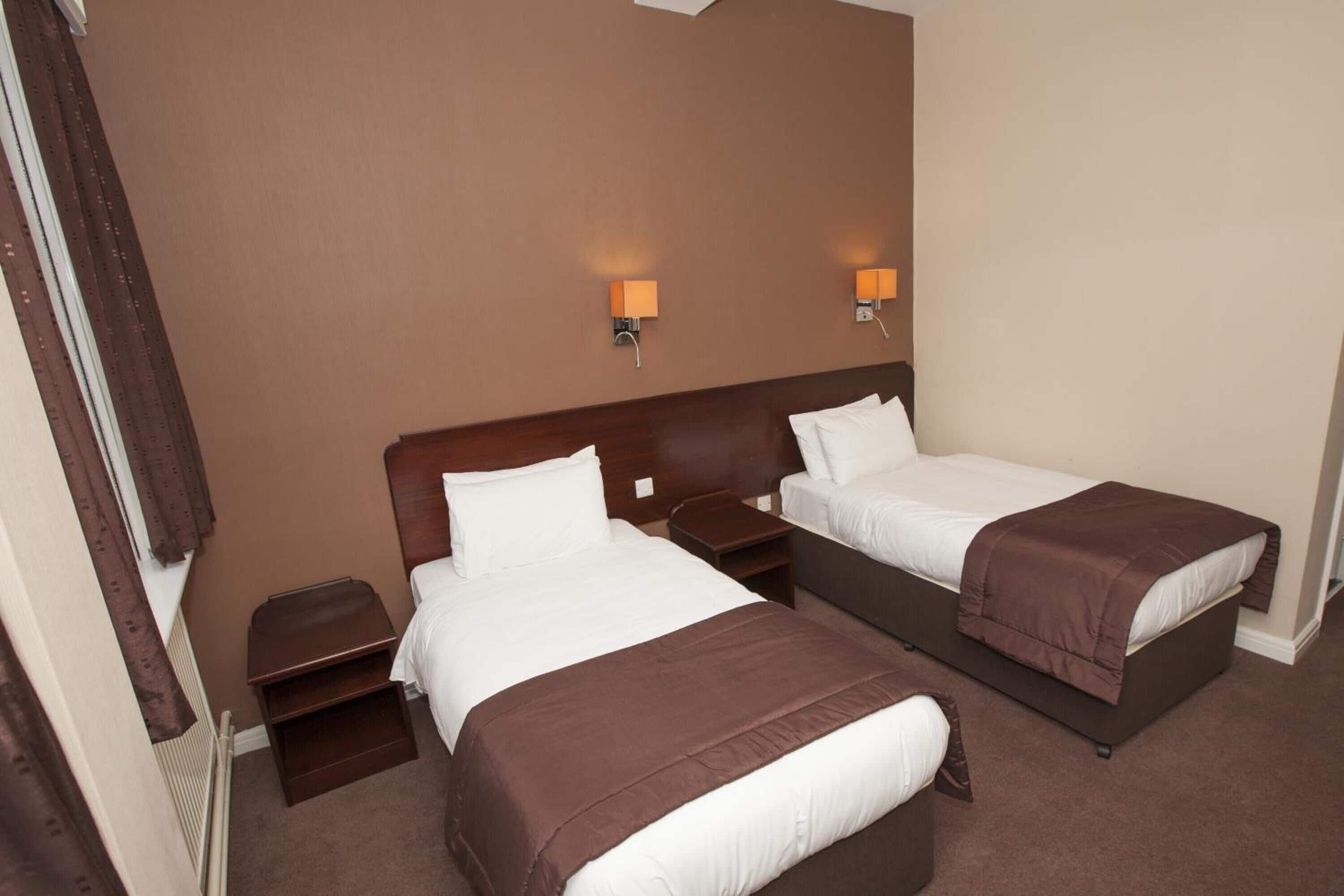 Quarto de hotel com duas camas, uma mesinha de cabeceira e duas luminárias.
