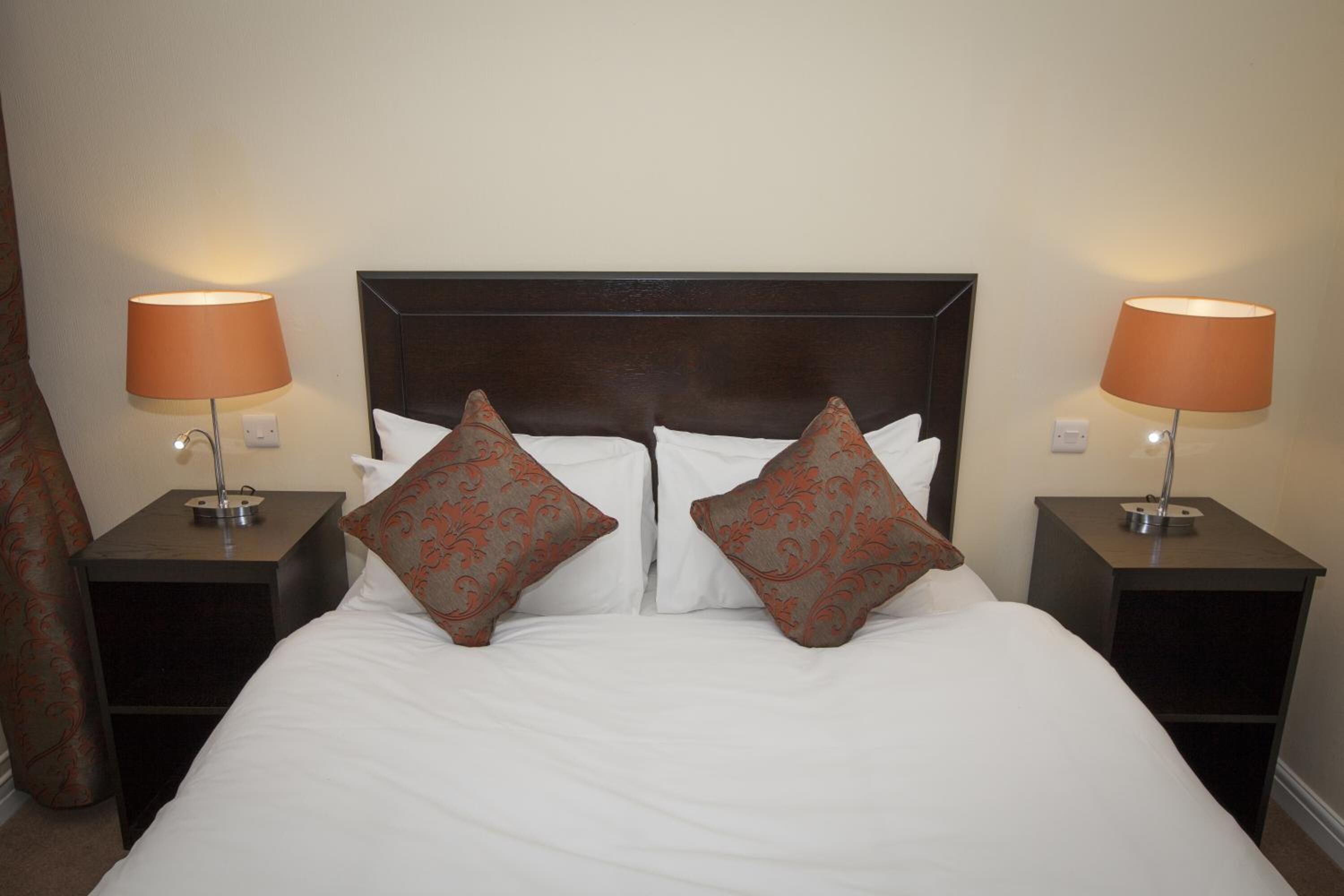 A neatly made bed with white linens and two decorative pillows, flanked by two bedside tables with lamps, in a simple, clean room.