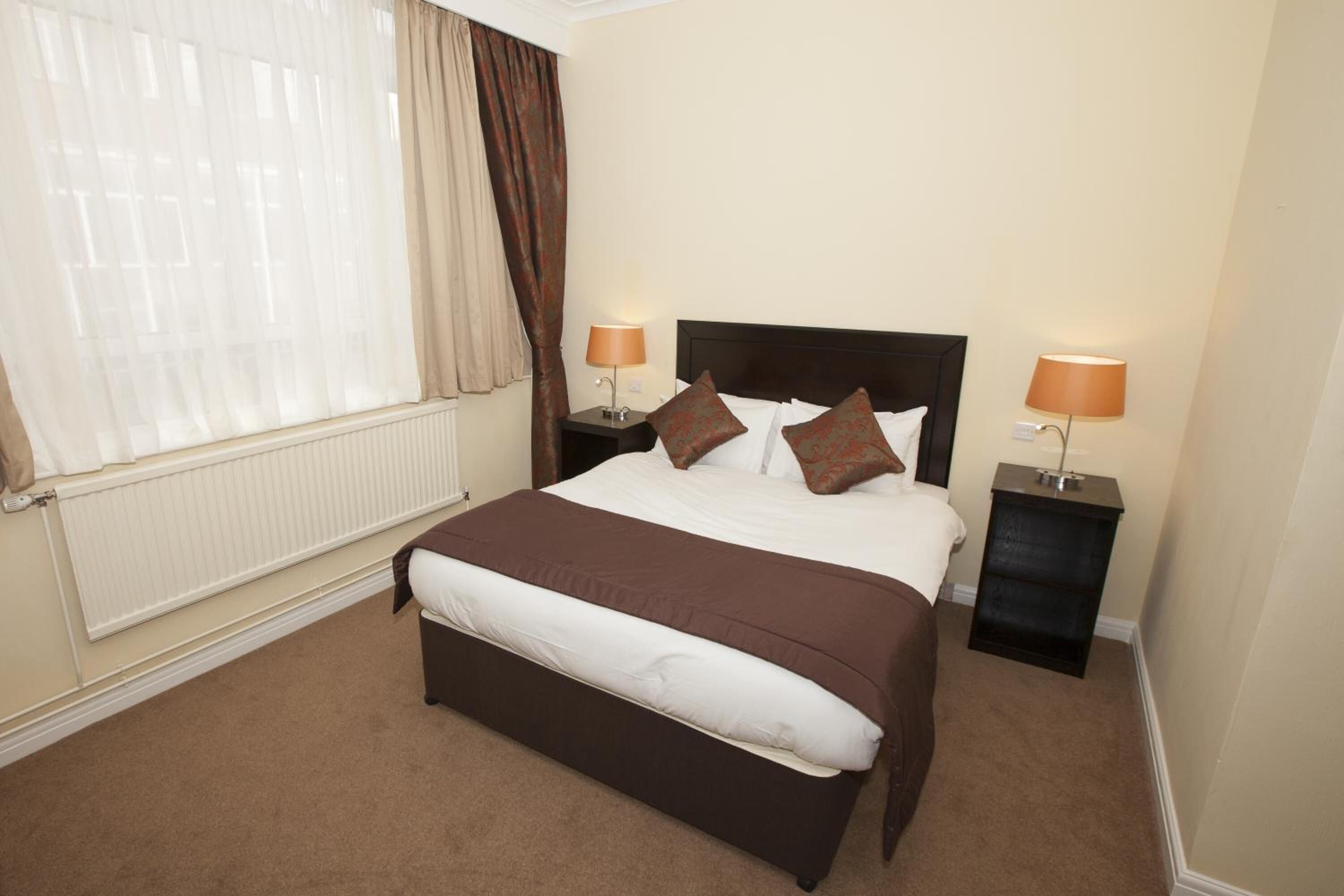A neatly made bed with a brown headboard, two bedside lamps, and a window with sheer curtains.