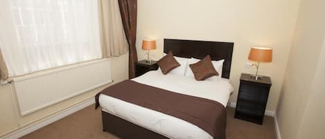A neatly made bed with a brown headboard, two bedside lamps, and a window with sheer curtains.