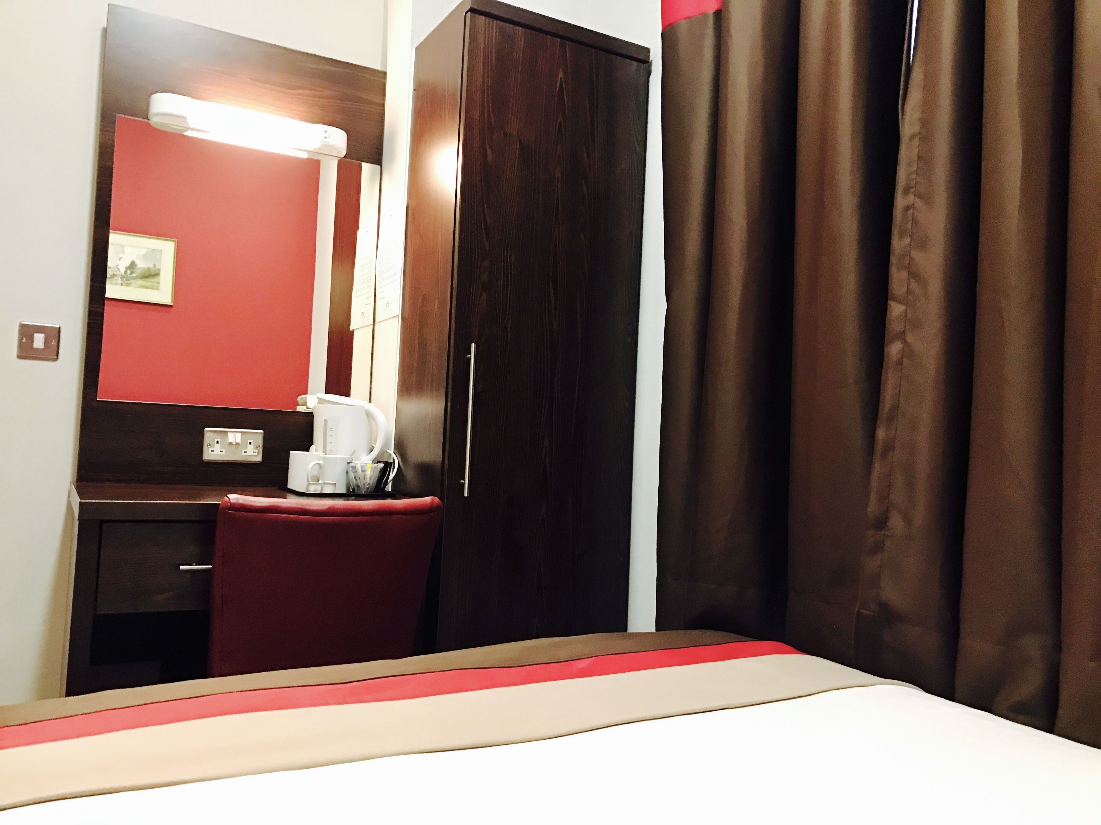 Habitación de hotel con cama, escritorio, silla, armario, espejo, cafetera y cortina.