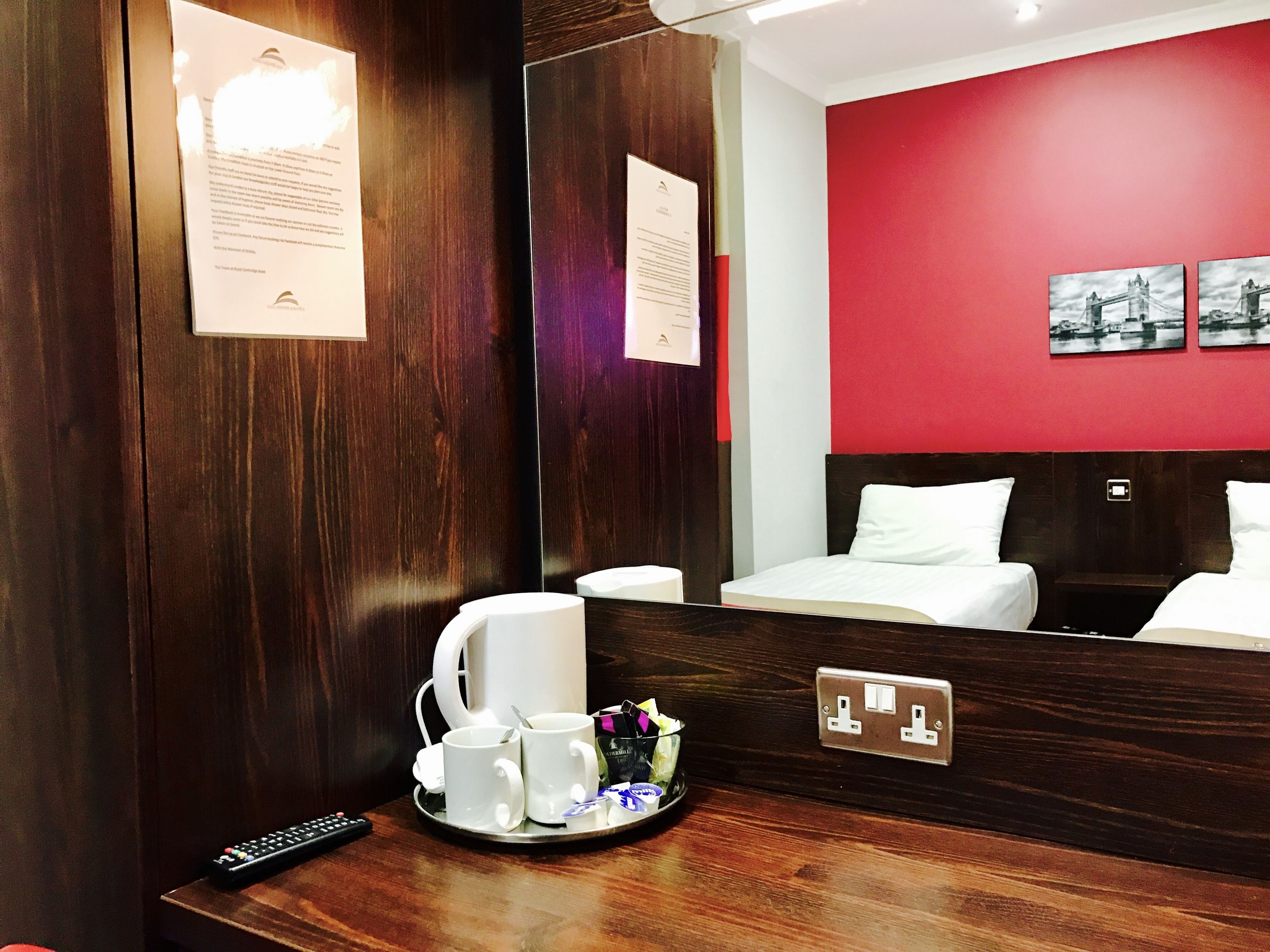A hotel room with a wooden headboard, a large mirror, a tray with two mugs, a remote control, and a small table with a bottle and a glass.