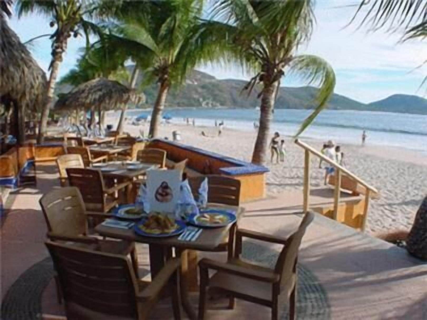 Un restaurante frente a la playa con terraza, palmeras y vistas al mar.