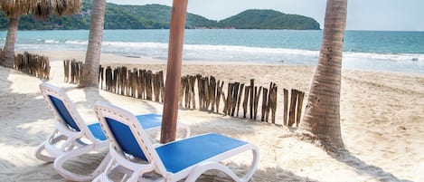 A beach with two white lounge chairs with blue cushions, a thatched umbrella, and a wooden fence.
