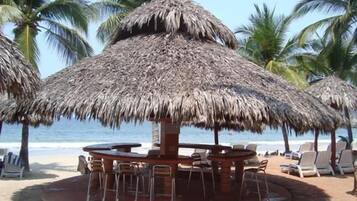 A beachside restaurant with thatched umbrellas, outdoor seating, and a view of the ocean.